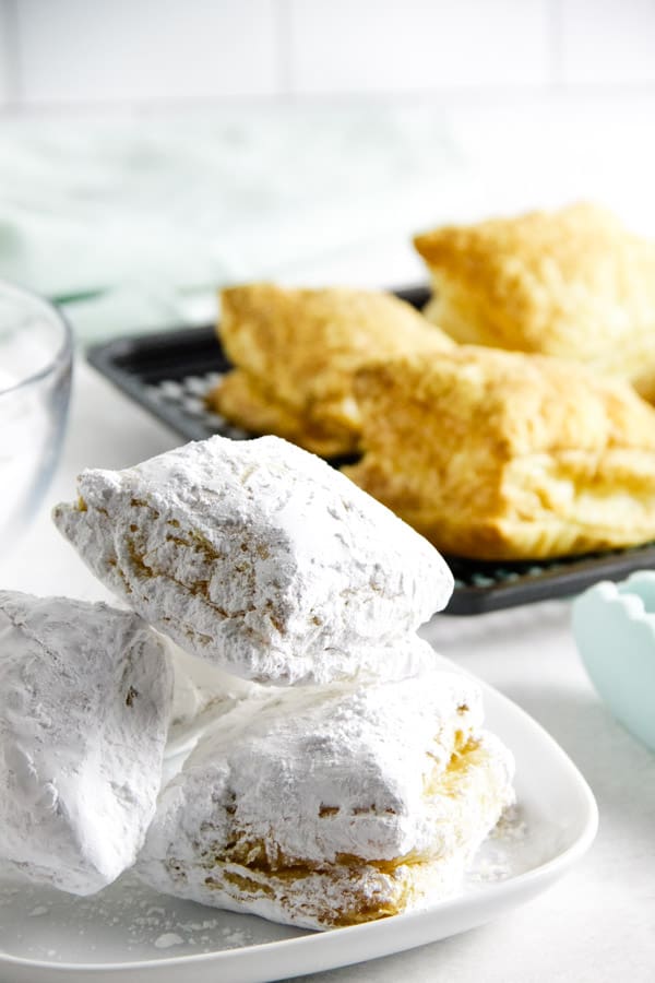 Easy 2 ingredient Air Fryer Beignets! Made with buttery puff pastry and coated with powdered sugar with no oil! Ready in 9 minutes! Air Fryer Beignets on white plate with black tray in background.