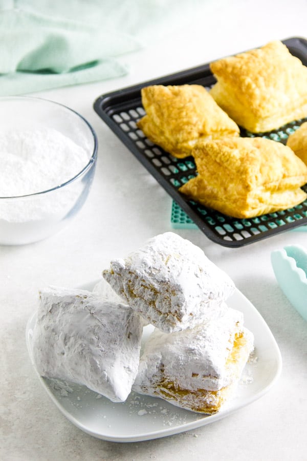Easy 2 ingredient Air Fryer Beignets! Made with buttery puff pastry and coated with powdered sugar with no oil! Ready in 9 minutes! Air Fryer Beignets on white plate.