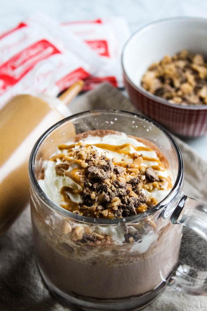 Peanut Butter Hot Chocolate in glass mug with toppings in background.